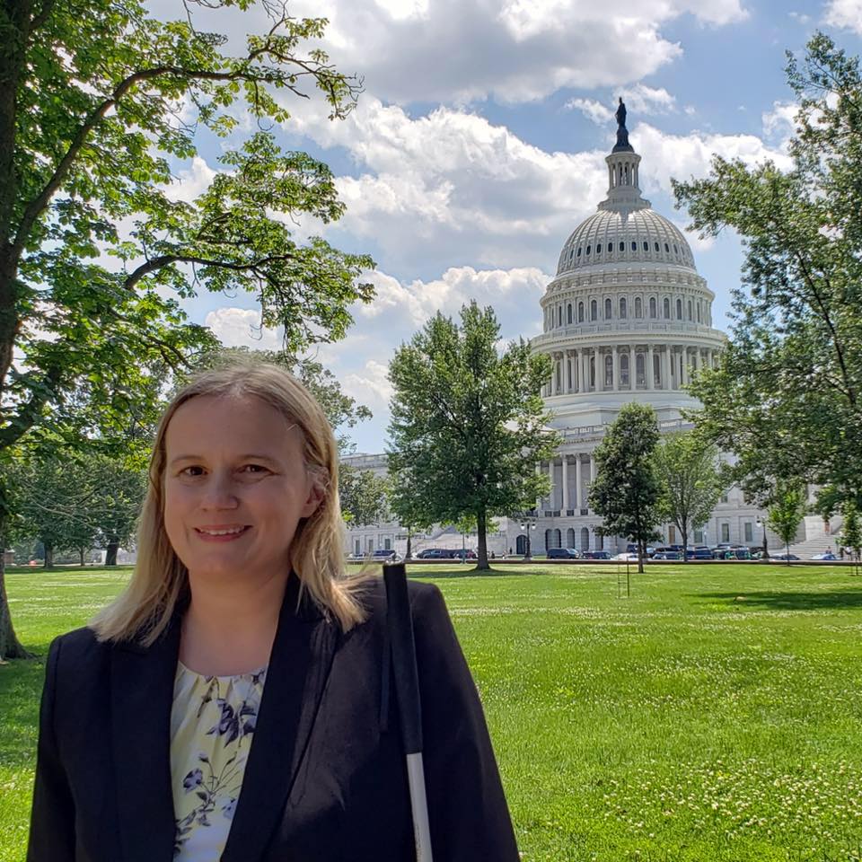 A woman with dark blonde hair stands in front of the US Capitol Building. She is wearing a black business suit. A long white cane is at her side.